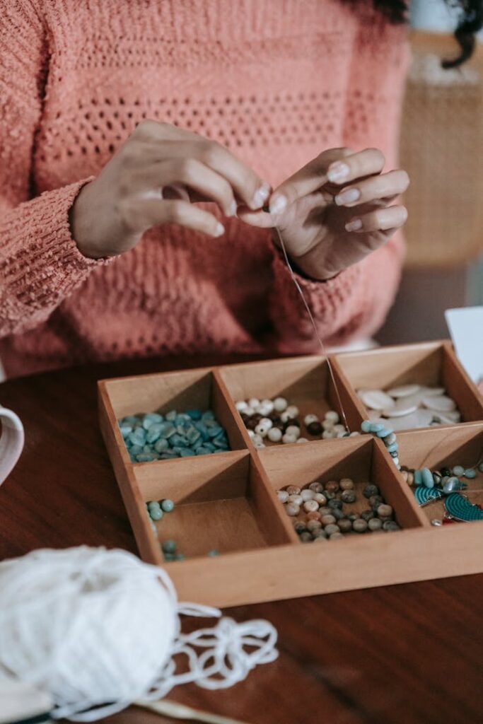 Crop unrecognizable ethnic female in light orange sweater doing beading with colorful beads and fishing line